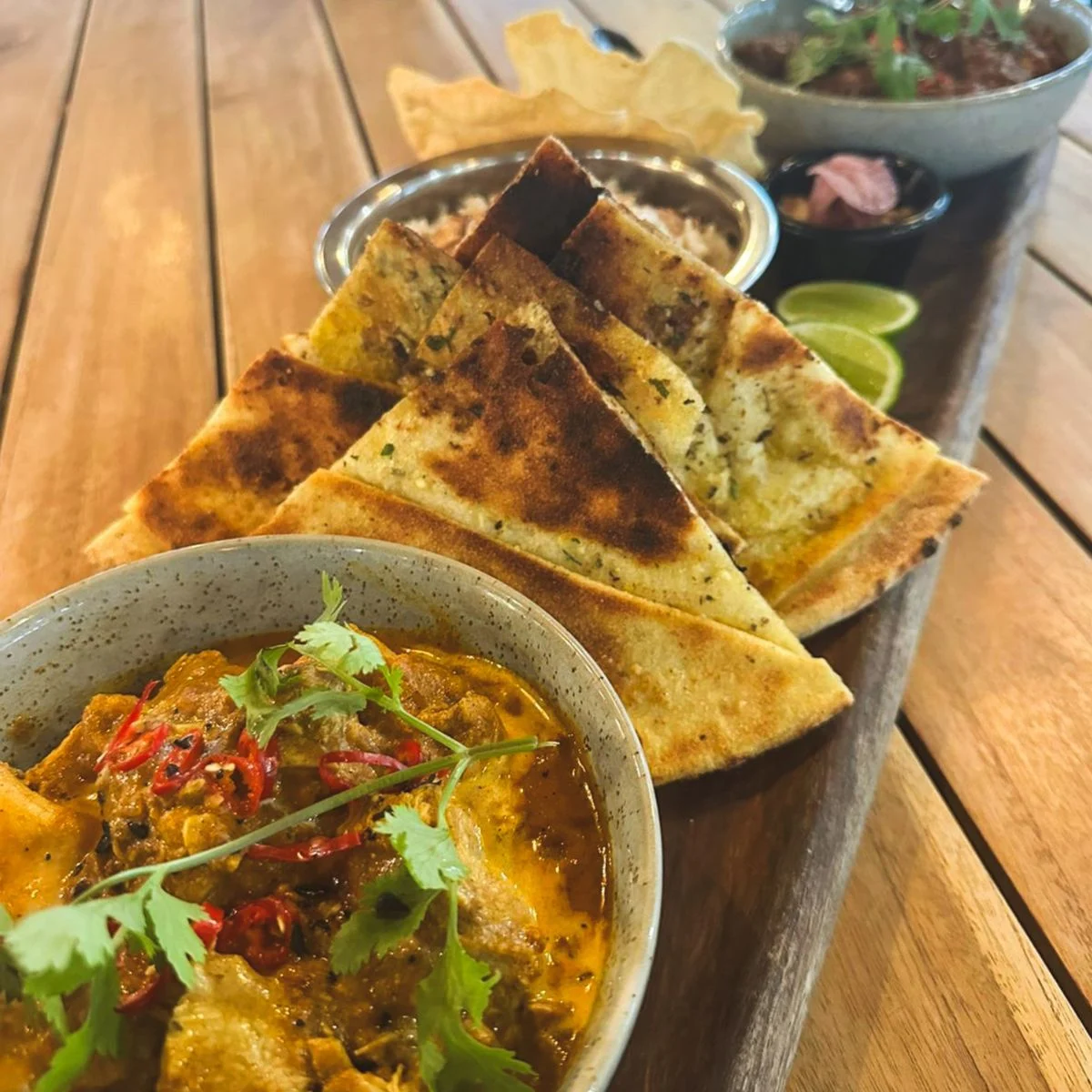 A wooden platter with sliced naan bread, a bowl of curry garnished with cilantro and red chili, rice, lime wedges, papadum, and another small bowl, all arranged on a wooden table.