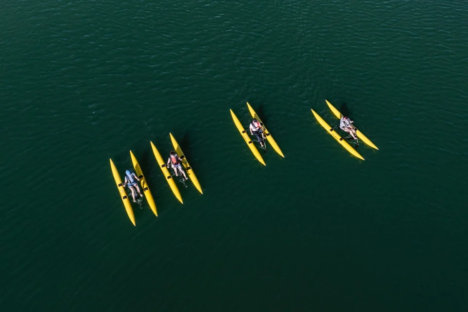 Four people riding yellow waterbikes in pairs on calm, dark green water. The kayaks are bright yellow, creating a striking contrast with the water. The scene is captured from above, showing two pairs of kayakers close together and one individual slightly apart.