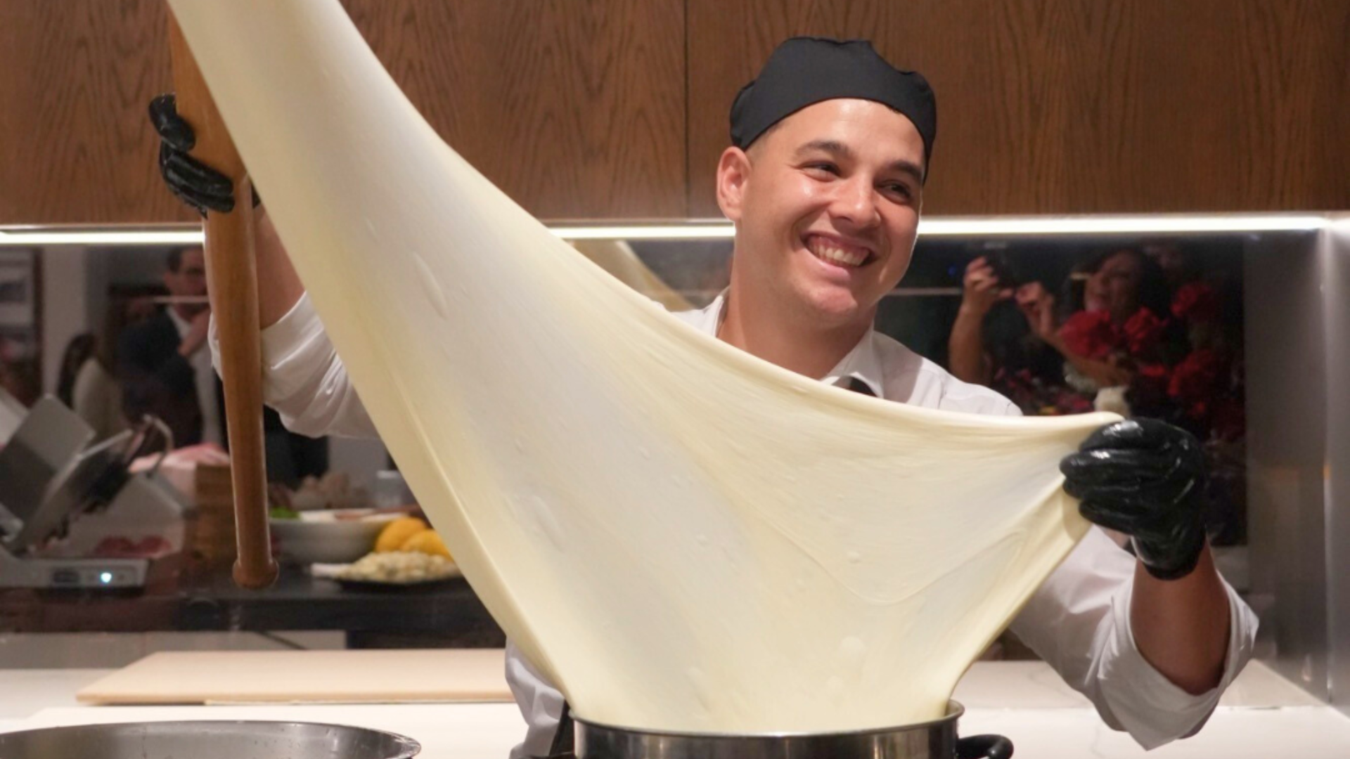 A smiling chef in a black hat and gloves stretches a large, thin sheet of dough in a kitchen, holding it wide above a pot with both hands. A wooden cabinet and people are visible in the background.