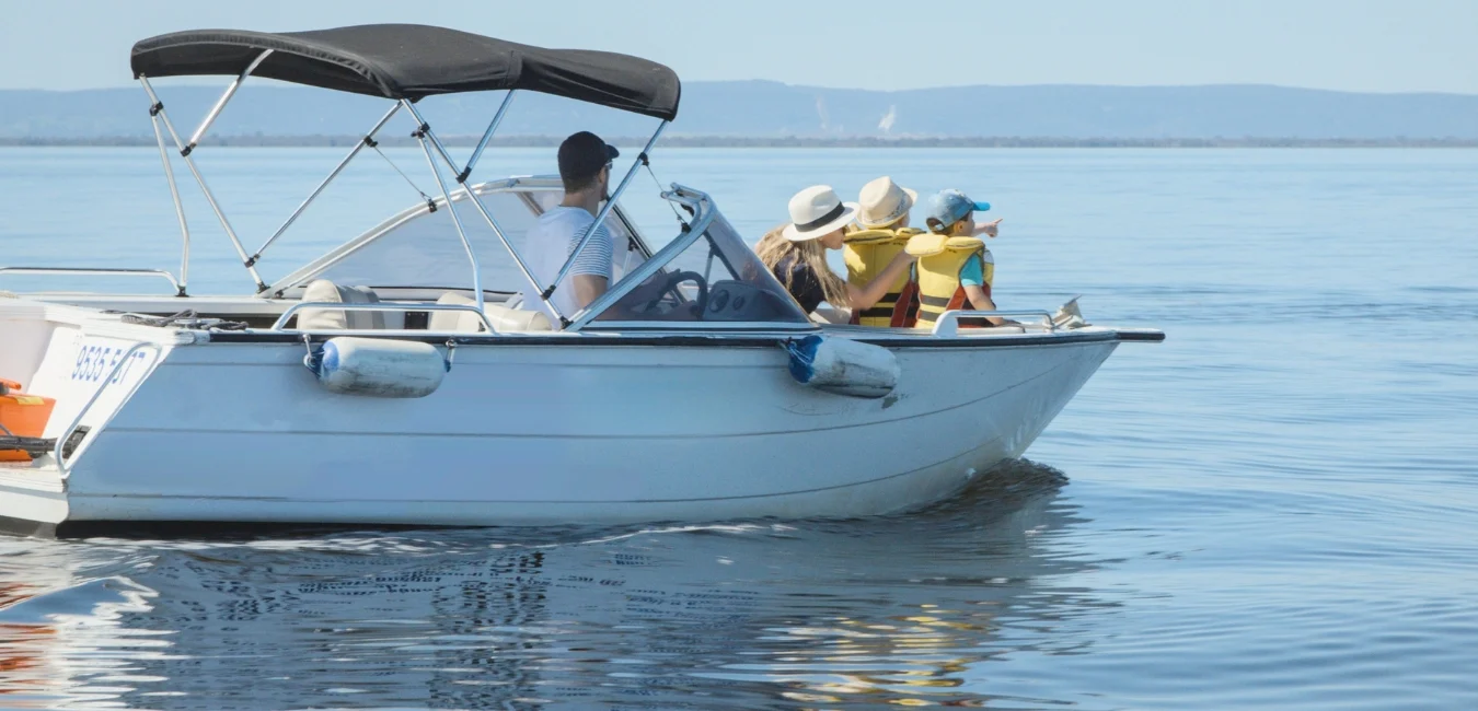 A small motorboat with a black canopy on a calm lake. A man steers while three people wearing life jackets and sun hats sit on the bow, enjoying the view. The distant shoreline is faintly visible under a clear blue sky.