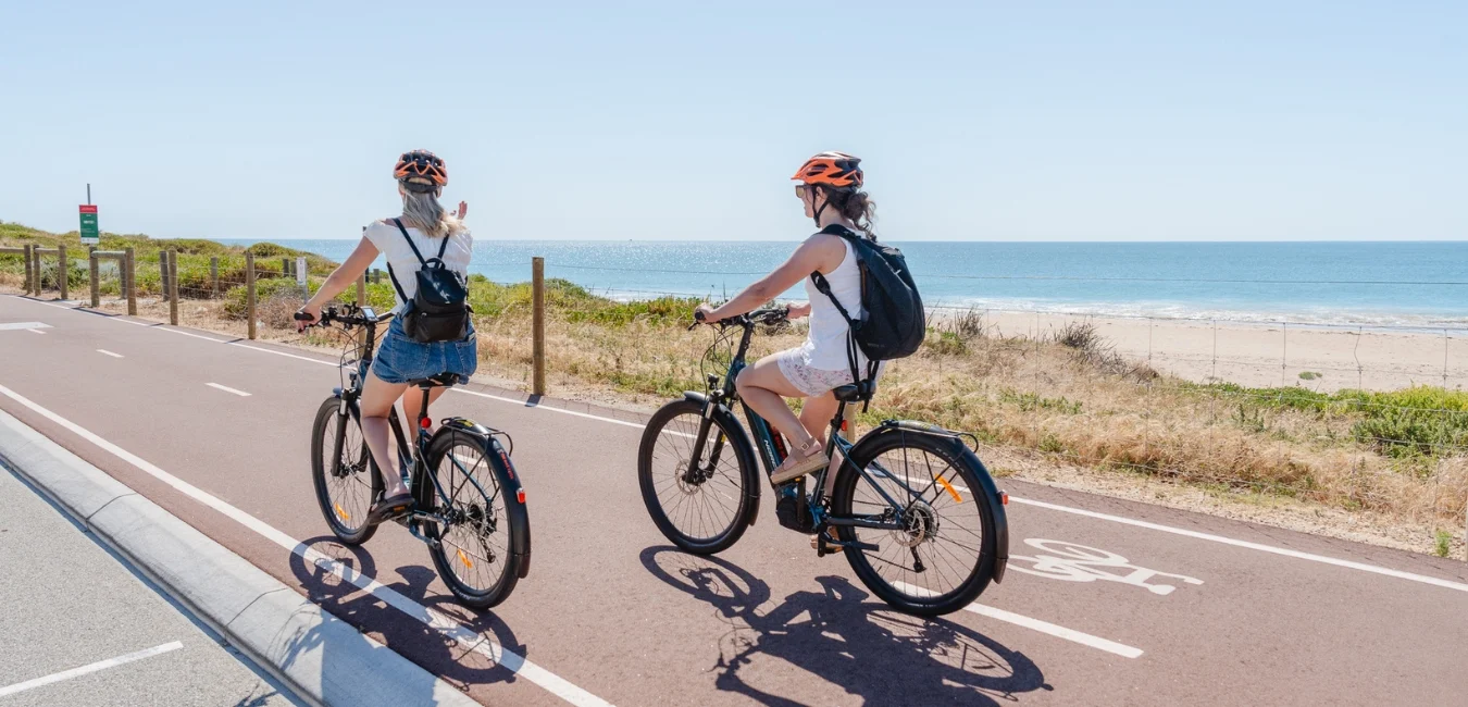 Two people wearing helmets and backpacks ride bicycles along a coastal bike path. The path runs parallel to a sandy beach and a blue ocean under a clear sky. One cyclist wears a gray top and shorts, the other a white dress.