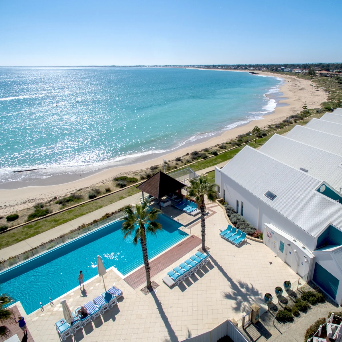 Aerial view of a seaside resort with a pool, lounge chairs, palm trees, and white buildings next to a sandy beach and turquoise ocean under a clear blue sky.