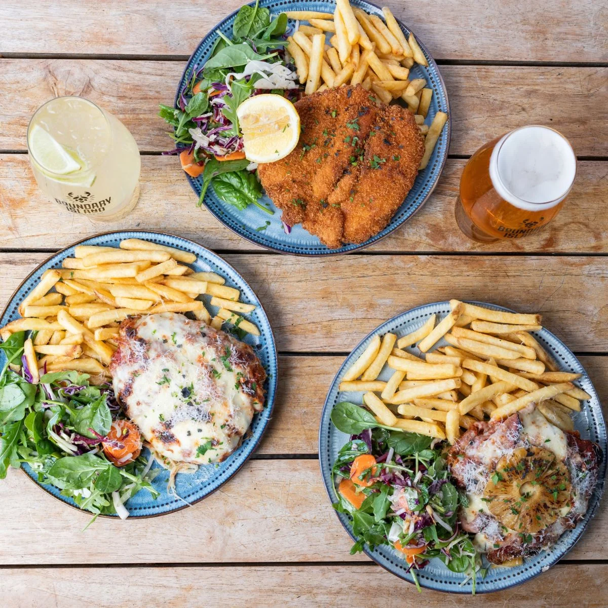 Three plates of pub-style meals on a wooden table, each with fries, salad, and chicken parmigiana, alongside a glass of beer and a glass of lemon drink.