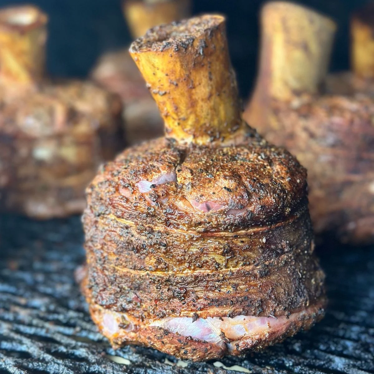 Close-up of a seasoned, smoked tomahawk-style cut of meat with a bone sticking out, resting on a dark grill surface. The meat appears juicy and well-cooked, with visible spices and a charred exterior.