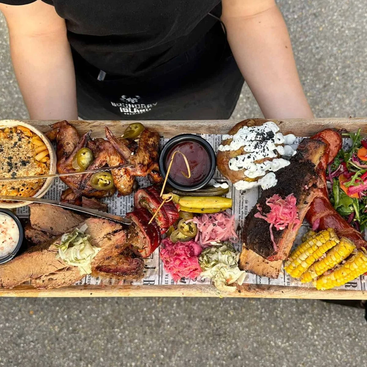 A person holds a wooden tray filled with assorted barbecue meats, corn on the cob, wings, pickles, salad, sauces, and bread..