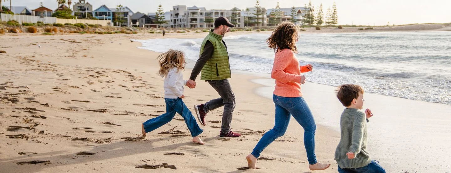 A man, woman, and two children run barefoot along a sandy beach near the ocean, with waves approaching and buildings visible in the background.