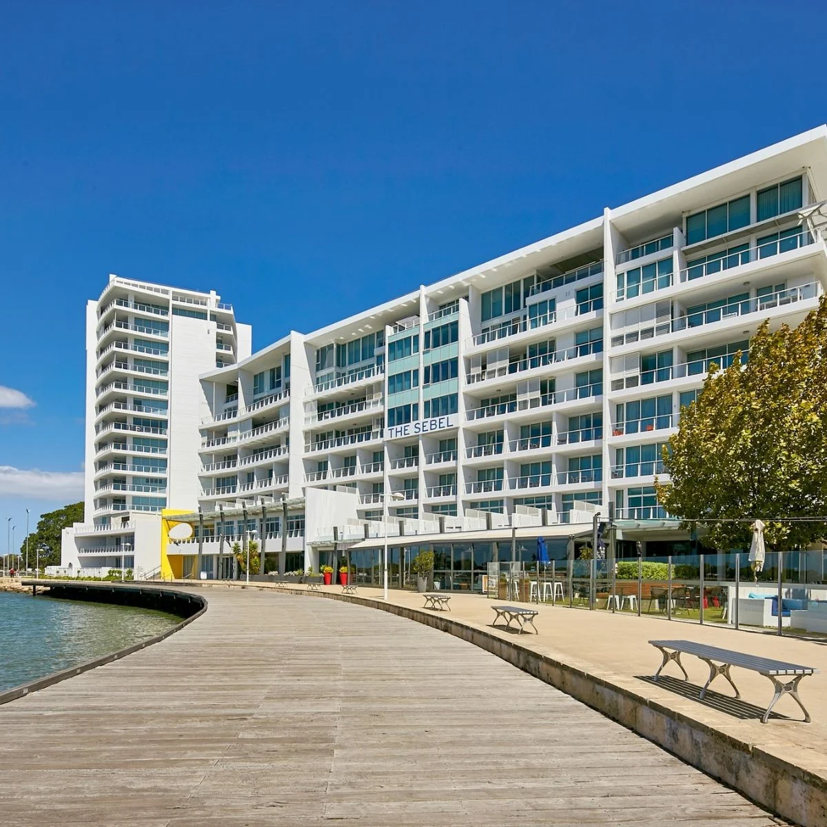 A modern white hotel with glass balconies overlooks a wooden boardwalk and calm waterfront under a clear blue sky. Benches and trees line the walkway, creating a relaxing, inviting atmosphere.