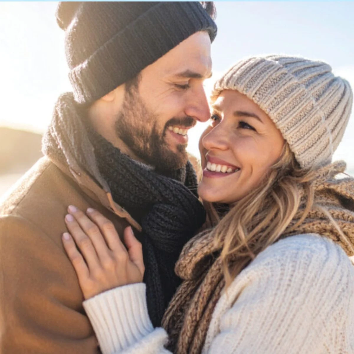 A smiling couple dressed in warm winter clothing, including knit hats and scarves, stands close together outdoors on a sunny day, looking at each other affectionately.