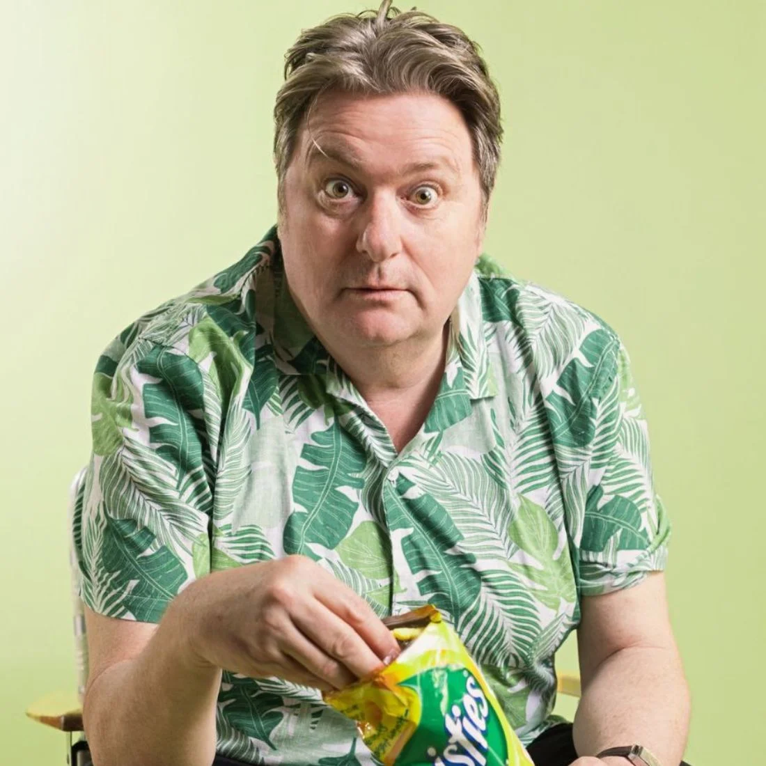A surprised man in a green leaf-patterned shirt looks at the camera while holding an open bag of crisps against a light green background.