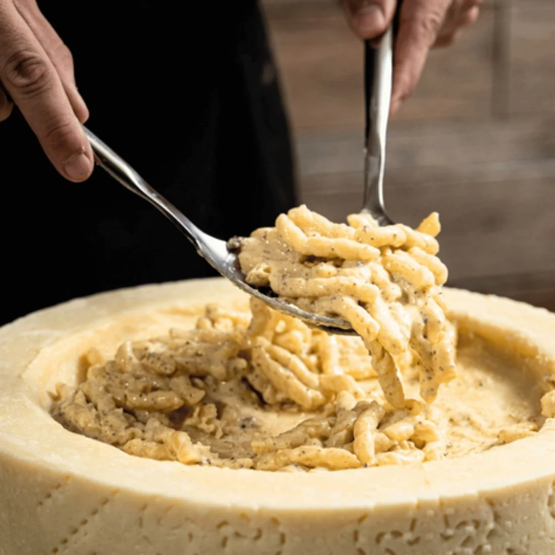 A person serves creamy pasta with tongs from a hollowed-out wheel of cheese, mixing the pasta inside the cheese for extra flavor.