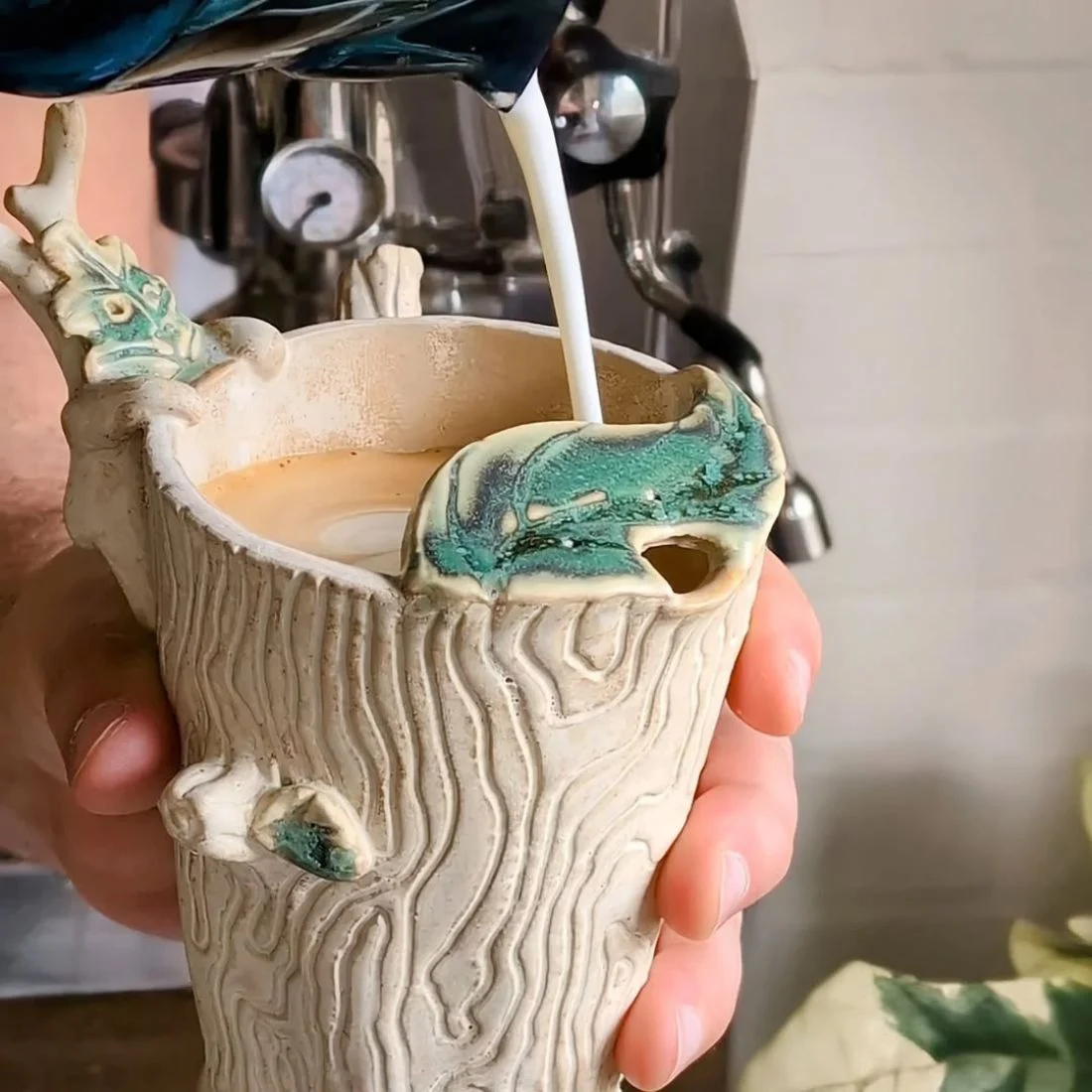A hand holds a ceramic mug designed to look like a tree trunk with green accents while milk is being poured into coffee from above. An espresso machine is visible in the background.