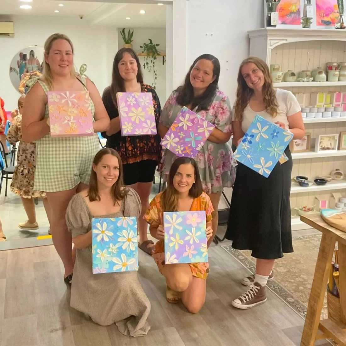 Six smiling women stand and kneel indoors, each holding a colorful canvas with painted flowers. The background has shelves with ceramics and art supplies, giving a cozy, creative studio atmosphere.