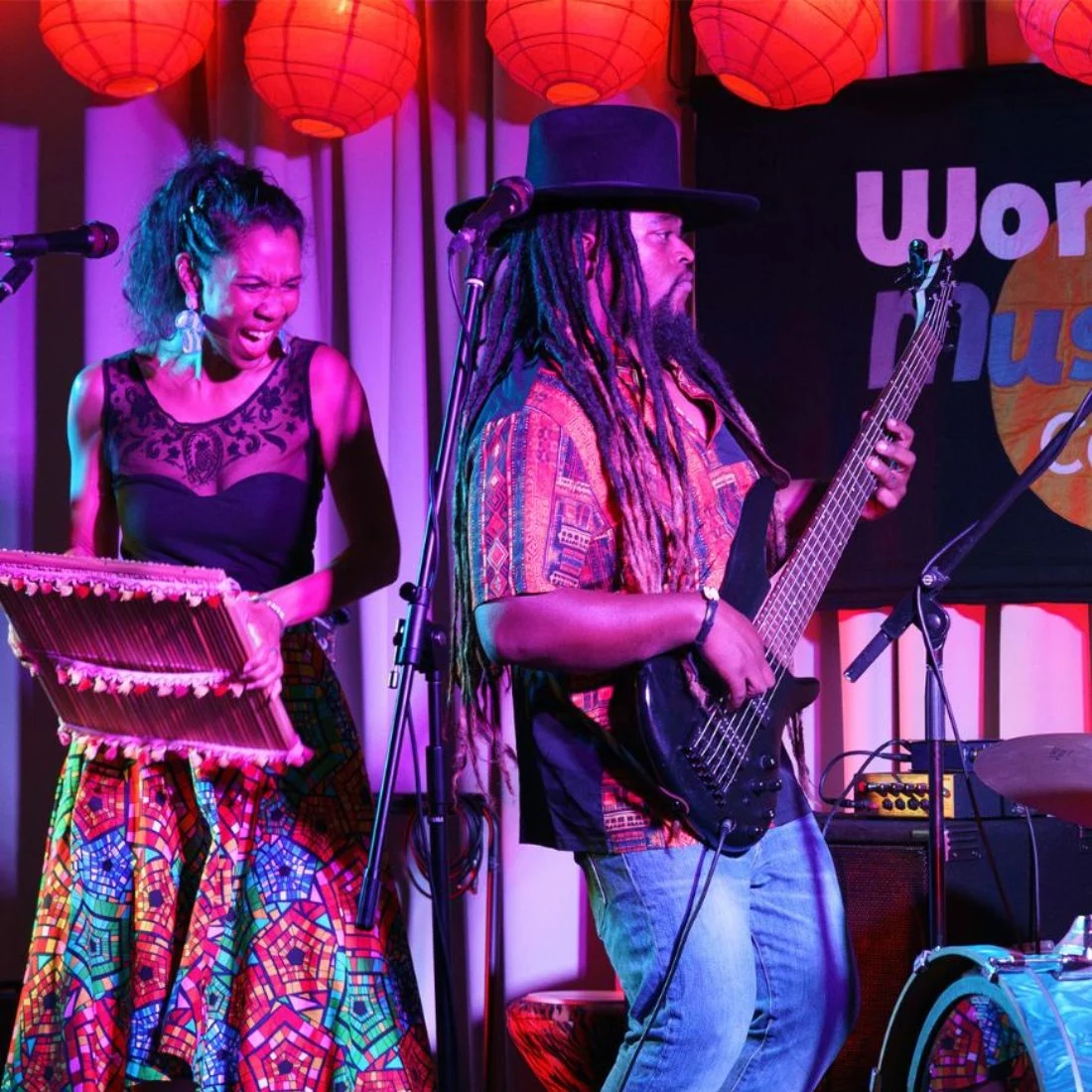 A woman plays a percussion instrument and smiles, while a man with long dreadlocks and a hat plays an electric bass guitar onstage. They are lit by colorful lights and red lanterns, with a “World Music” banner behind them.