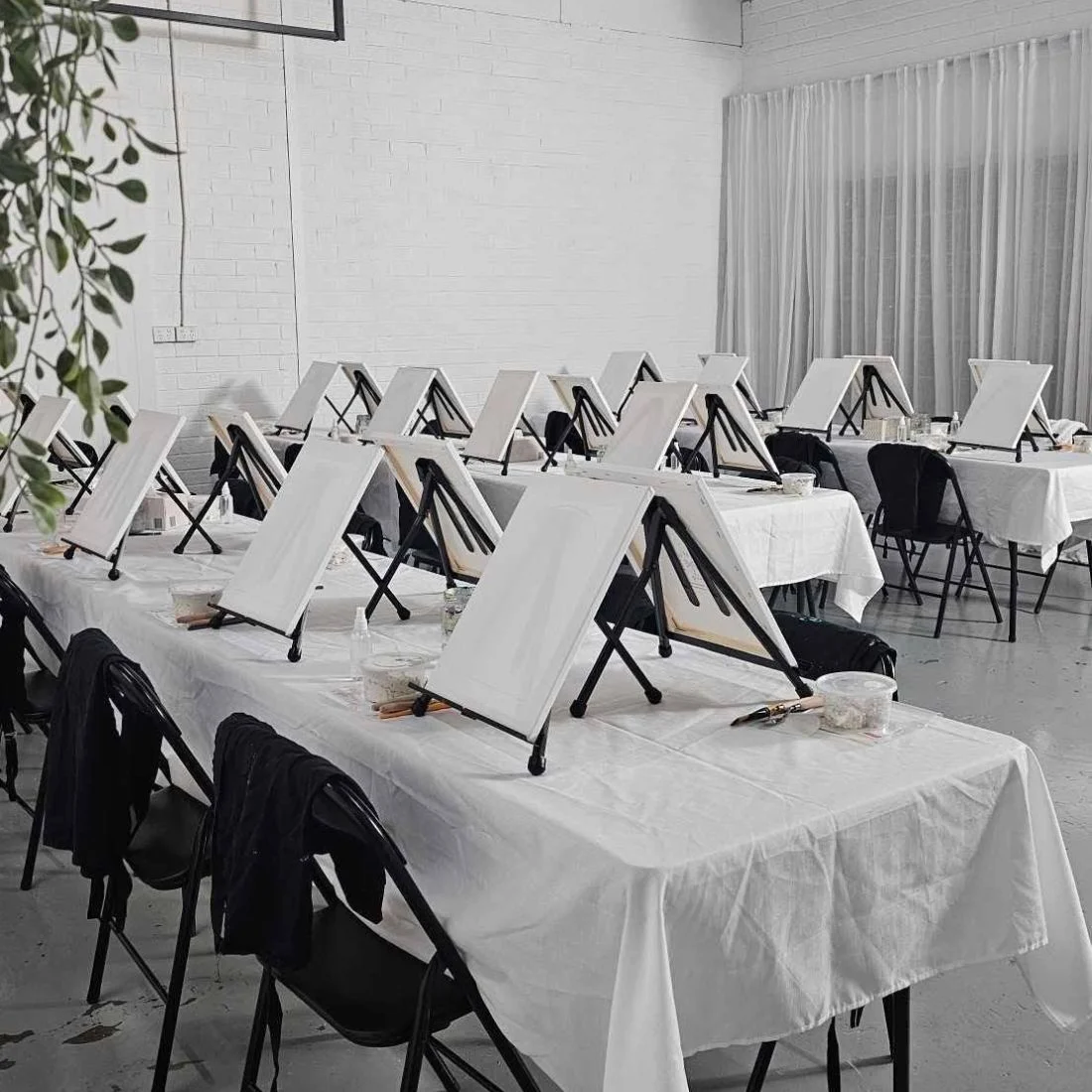 A studio with rows of tables covered in white cloth, each set with a blank canvas on an easel, paint supplies, and black chairs, ready for a group painting session.