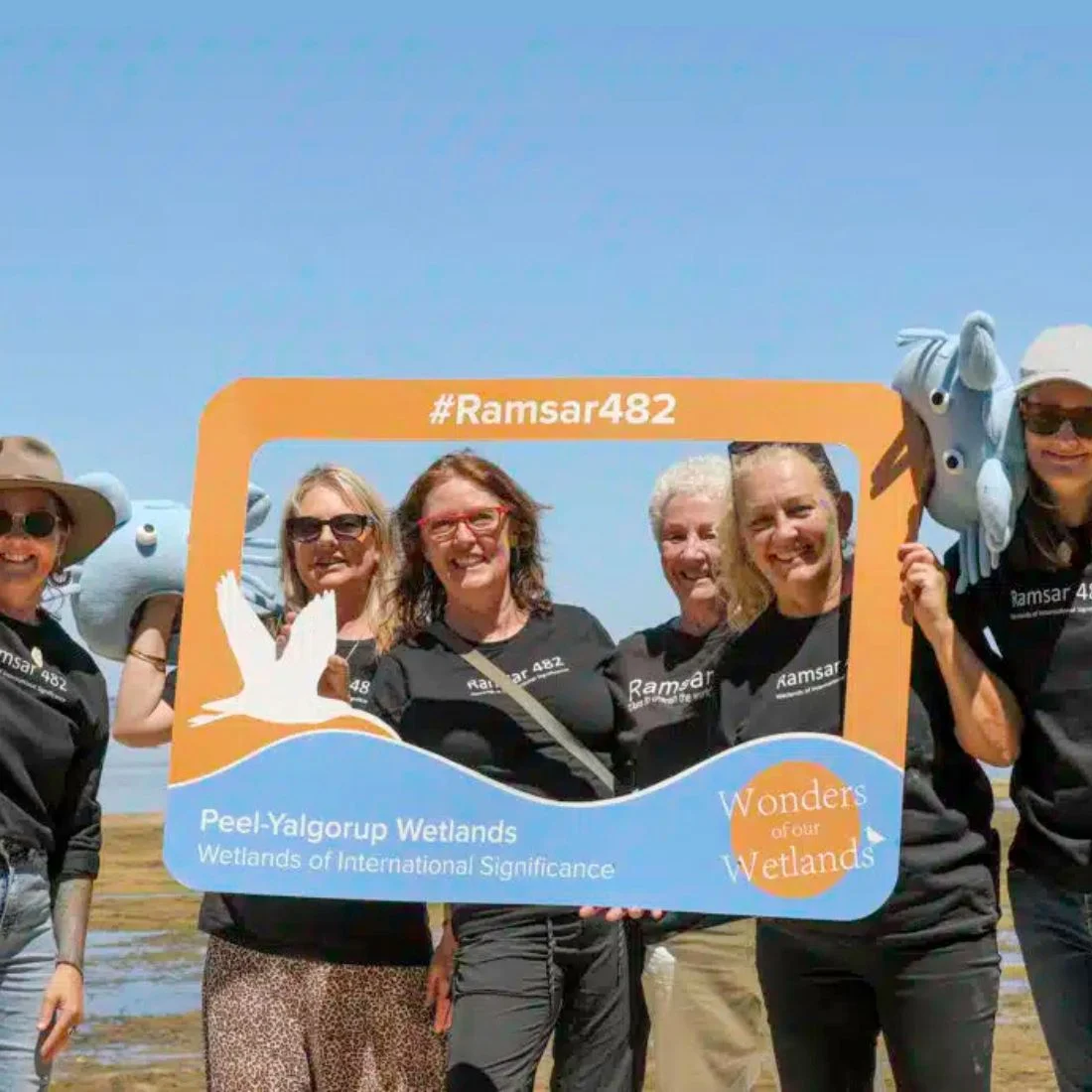A group of six smiling people in black shirts hold a large photo frame prop labeled #Ramsar482 and Peel-Yalgorup Wetlands, with a bright blue sky in the background. Two hold plush animal toys.