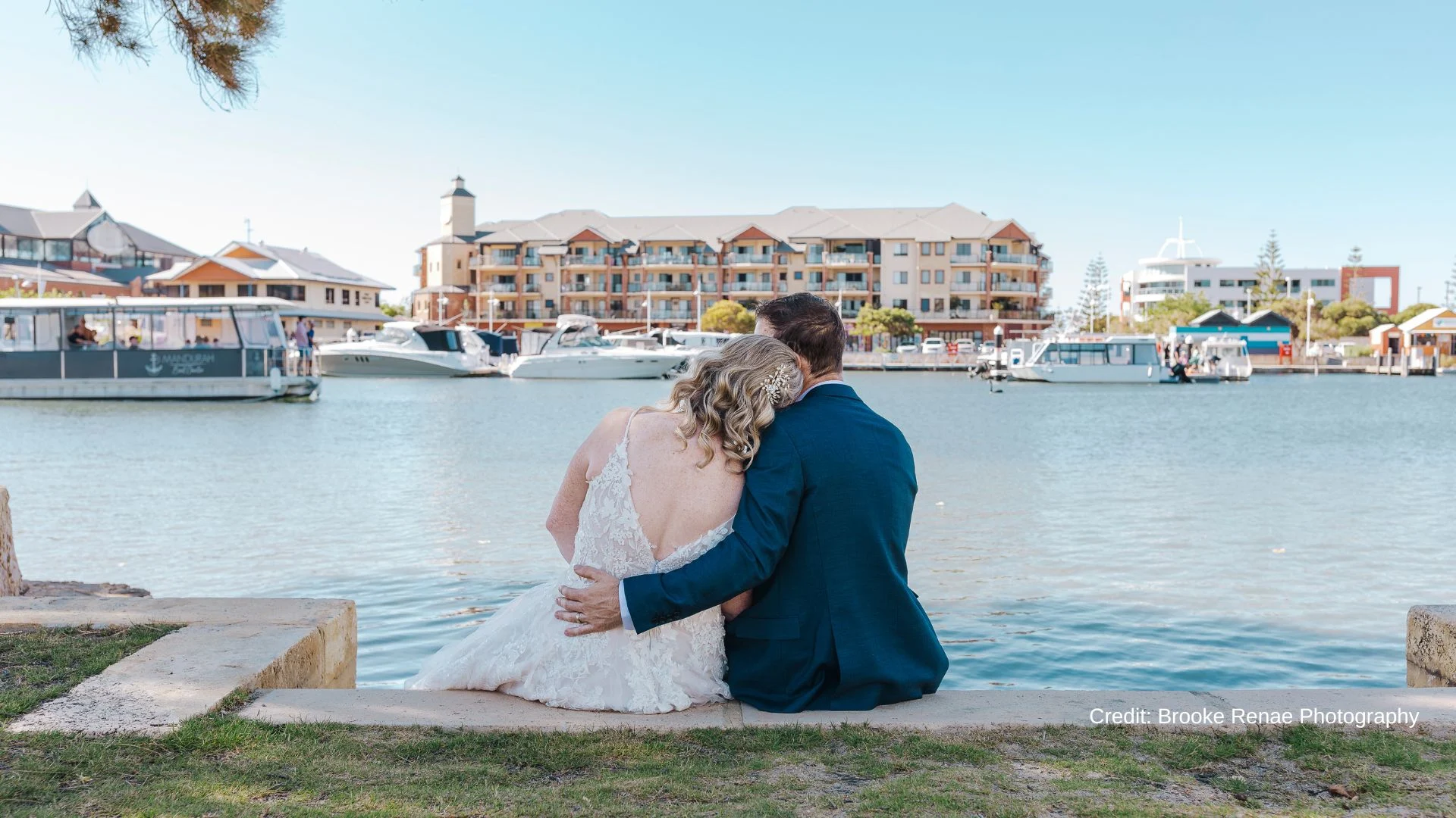 A bride in a white dress and a groom in a blue suit sit on a stone edge by the water, embracing and looking at boats and waterfront buildings under a clear blue sky.