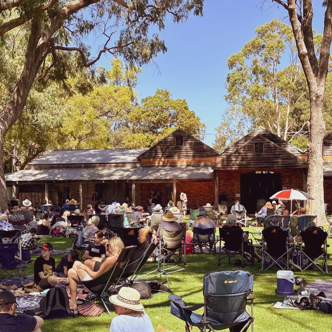 People sit on lawn chairs and blankets in a grassy area under trees, enjoying a sunny day in front of rustic wooden buildings. Some wear hats or sunglasses, and the atmosphere is relaxed and leisurely.