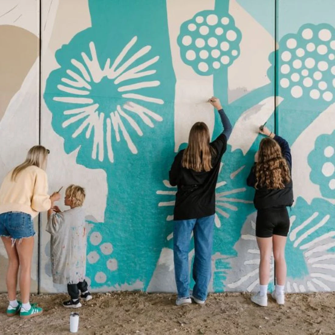 Four people, including two children and two adults, are painting a colorful mural with abstract white floral patterns on a large outdoor wall. They are focused on their work, and painting supplies are on the ground nearby.